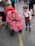 A baby in a stroller wears a bright red dress with white polka dots and a matching red headband featuring a flower. Beside the stroller, a young boy dressed in vintage-style clothing with a flat cap, white shirt, red suspenders, and striped pants looks on. The scene is set on a street or public area with various people and stalls in the background.