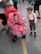 A baby in a stroller wears a bright red dress with white polka dots and a matching red headband featuring a flower. Beside the stroller, a young boy dressed in vintage-style clothing with a flat cap, white shirt, red suspenders, and striped pants looks on. The scene is set on a street or public area with various people and stalls in the background.