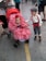 A baby in a stroller wears a bright red dress with white polka dots and a matching red headband featuring a flower. Beside the stroller, a young boy dressed in vintage-style clothing with a flat cap, white shirt, red suspenders, and striped pants looks on. The scene is set on a street or public area with various people and stalls in the background.