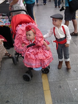 A baby in a stroller wears a bright red dress with white polka dots and a matching red headband featuring a flower. Beside the stroller, a young boy dressed in vintage-style clothing with a flat cap, white shirt, red suspenders, and striped pants looks on. The scene is set on a street or public area with various people and stalls in the background.