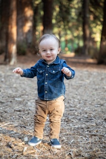 A young child with light hair stands confidently in a wooded area, wearing a denim jacket, brown pants, and navy slip-on shoes. The background features tall trees and a path covered with dry leaves and twigs, suggesting an outdoor, forest setting.