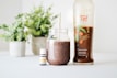 A clear glass jar filled with a brown smoothie and a straw is placed next to a bottle labeled 'Ningxia Red' and a small bottle of essential oil. The background features green potted plants, adding a fresh and natural aesthetic.