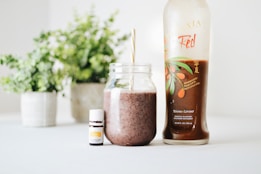 A clear glass jar filled with a brown smoothie and a straw is placed next to a bottle labeled 'Ningxia Red' and a small bottle of essential oil. The background features green potted plants, adding a fresh and natural aesthetic.