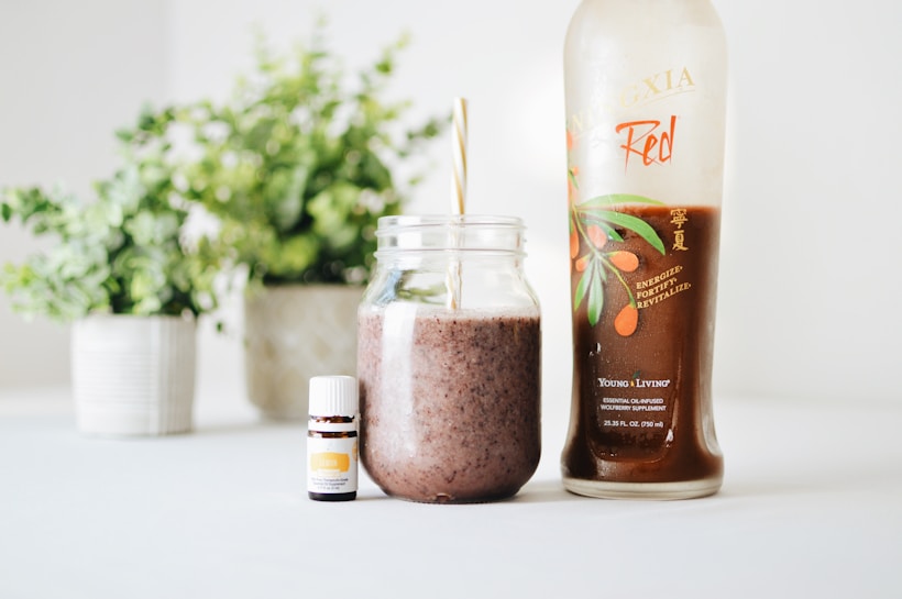 A clear glass jar filled with a brown smoothie and a straw is placed next to a bottle labeled 'Ningxia Red' and a small bottle of essential oil. The background features green potted plants, adding a fresh and natural aesthetic.