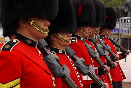 A group of uniformed soldiers wearing traditional red military jackets and black bearskin hats stand in a line, holding rifles. Each soldier has a chin strap decorated with gold chains. Their polished uniforms and disciplined formation reflect military precision. A metal railing and a statue are visible in the background, hinting at a ceremonial or official setting.