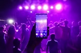 A lively party scene captured through the lens of a smartphone, with a crowd of people dancing under vibrant purple stage lights. The phone in the foreground mirrors the scene with its own display, emphasizing the integration of digital technology in social events.