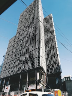 Tall, modern building with a grid-like facade under construction, set against a clear blue sky. The structure appears incomplete with visible steel frames and some signage at the ground level. Cars and construction materials are visible in the foreground, along with overhead wires.