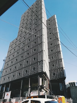 Tall, modern building with a grid-like facade under construction, set against a clear blue sky. The structure appears incomplete with visible steel frames and some signage at the ground level. Cars and construction materials are visible in the foreground, along with overhead wires.