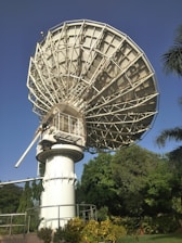A large satellite dish is positioned against a clear blue sky. The structure is metallic and intricate, towering over green foliage and well-maintained gardens. Palm trees and various bushes surround the base of the satellite.