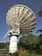A large satellite dish is positioned against a clear blue sky. The structure is metallic and intricate, towering over green foliage and well-maintained gardens. Palm trees and various bushes surround the base of the satellite.
