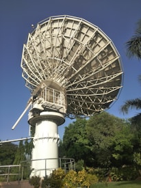 A large satellite dish is positioned against a clear blue sky. The structure is metallic and intricate, towering over green foliage and well-maintained gardens. Palm trees and various bushes surround the base of the satellite.