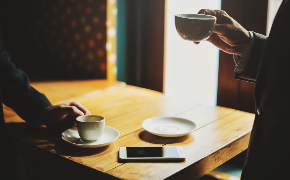 person holding white ceramic cup infront of person near the brown wooden table