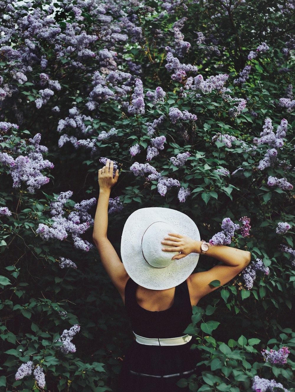 Femme portant un haut noir sans manches et tenant un chapeau et entouré de fleurs violettes