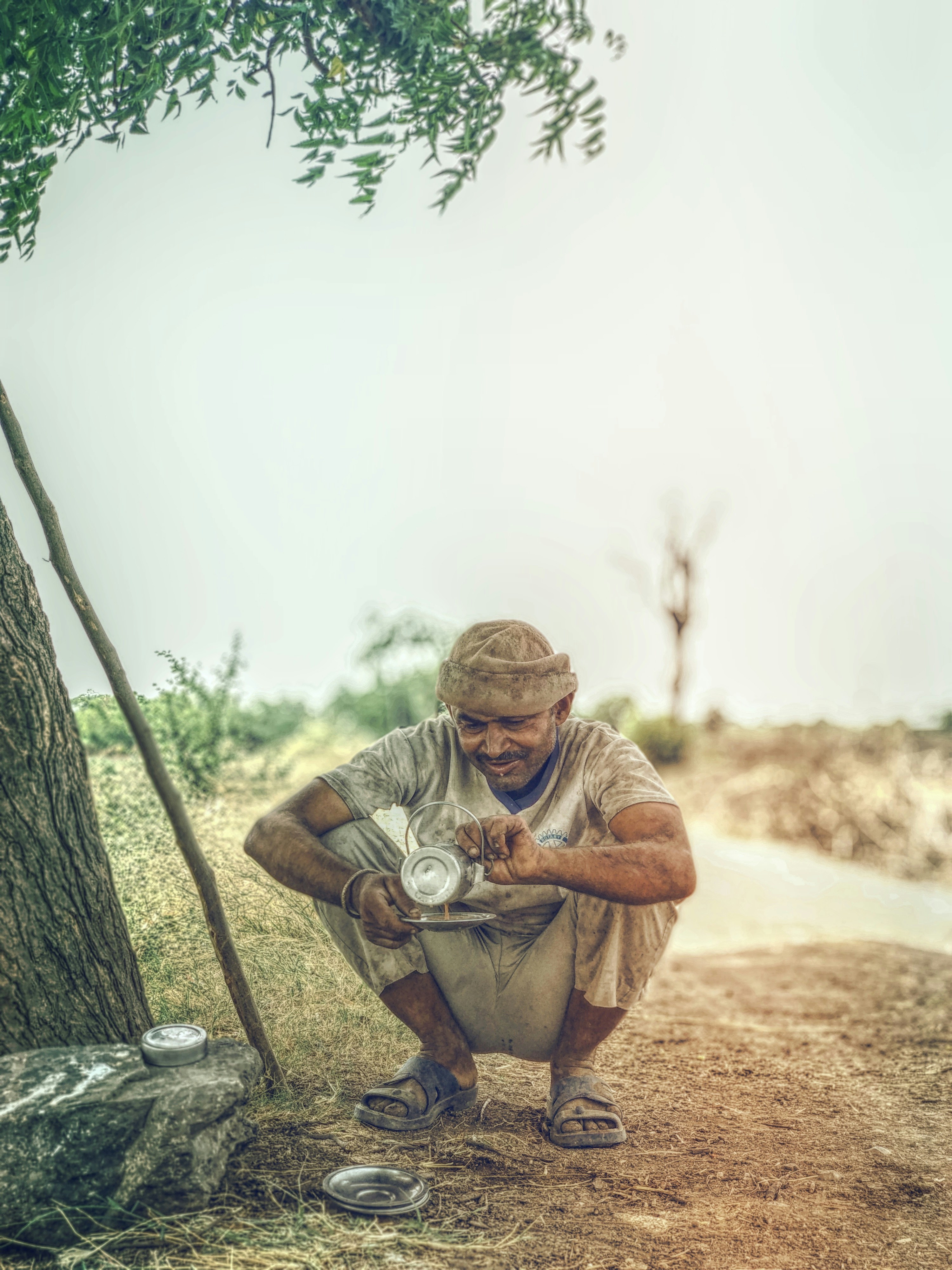 man pouring coffee on gray metal plate while crouching near tree during daytime