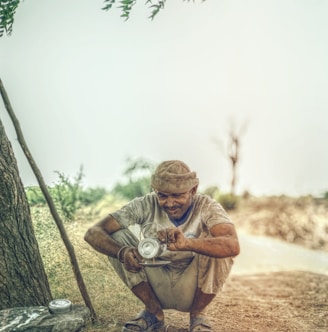 man pouring coffee on gray metal plate while crouching near tree during daytime