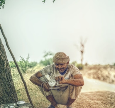 man pouring coffee on gray metal plate while crouching near tree during daytime