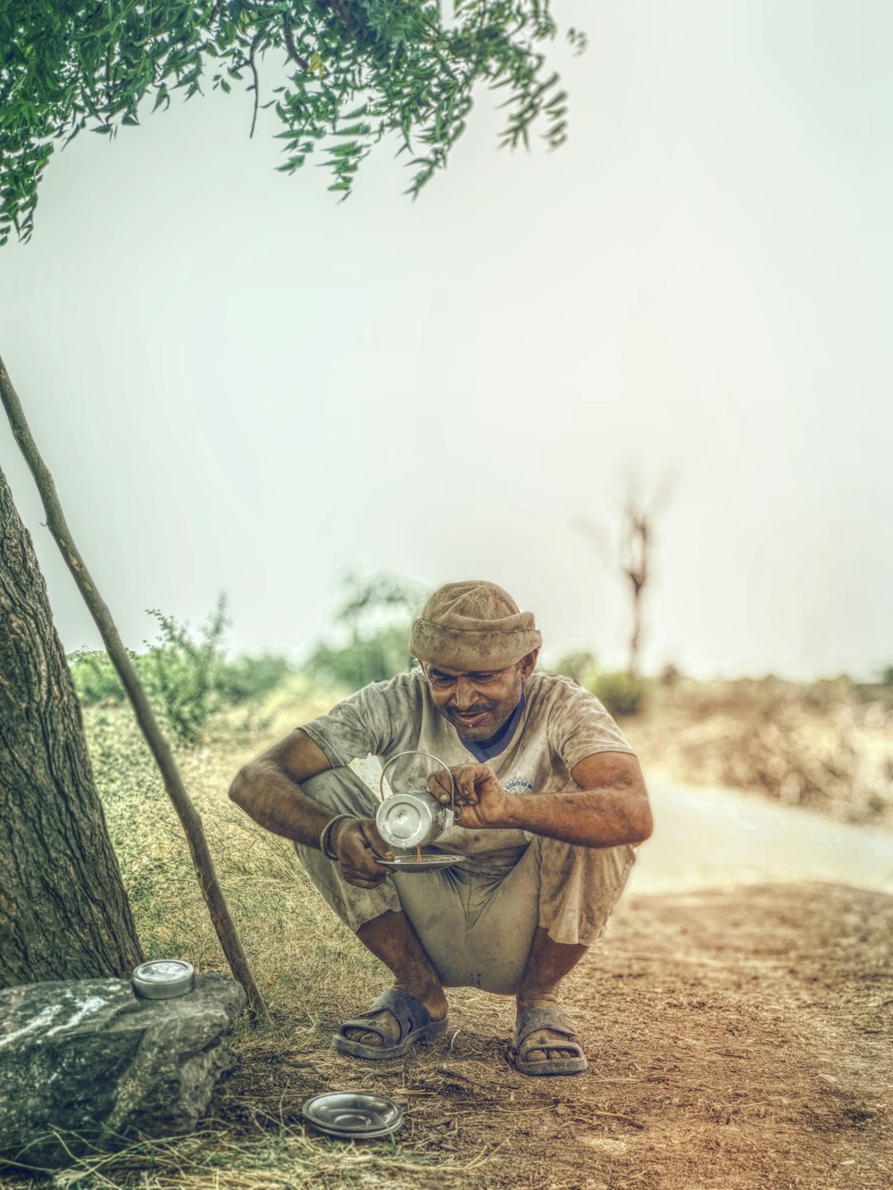 man pouring coffee on gray metal plate while crouching near tree during daytime