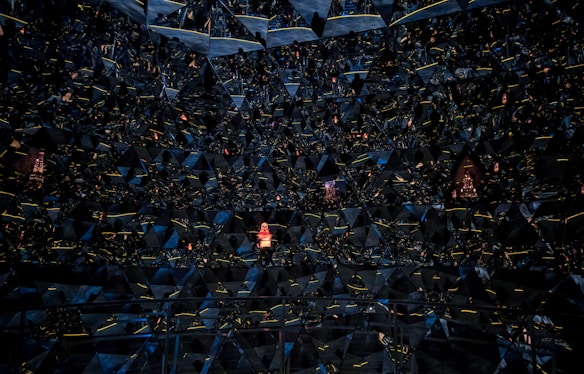 A complex pattern of mirrored triangular surfaces creating a kaleidoscopic effect. At the center, a small figure in a bright outfit stands out amidst the dark, glossy reflections. The geometric design is illuminated by subtle yellow and blue hues, with reflections creating an almost infinite depth.