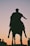 A silhouetted statue of a person on horseback stands against a gradient sky transitioning from a soft orange at the horizon to a gentle pink. The figure appears to be historic, likely a leader or warrior, with draped clothing visible in the silhouette.