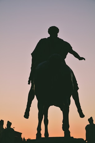 A silhouetted statue of a person on horseback stands against a gradient sky transitioning from a soft orange at the horizon to a gentle pink. The figure appears to be historic, likely a leader or warrior, with draped clothing visible in the silhouette.