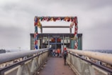 A walkway leads up to a structure with colorful cloth strips hanging across a metal frame at the top. The bridge-like walkway has metal sides and is elevated, with a view of a city skyline in the background. Several people walk towards the entrance of the structure, which appears to be an elevator or lift.