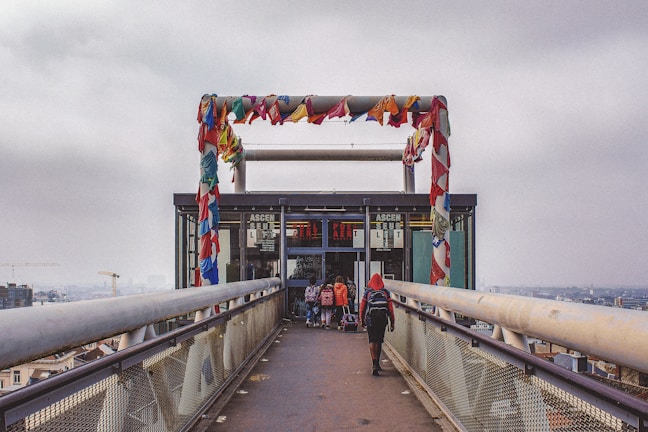 A walkway leads up to a structure with colorful cloth strips hanging across a metal frame at the top. The bridge-like walkway has metal sides and is elevated, with a view of a city skyline in the background. Several people walk towards the entrance of the structure, which appears to be an elevator or lift.