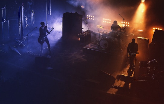 A live music performance with a band on stage, surrounded by a smoky atmosphere. The lighting is dramatic with warm spotlights illuminating the band members, creating a contrast with the dark, shadowy background.