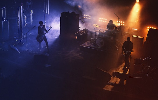 A live music performance with a band on stage, surrounded by a smoky atmosphere. The lighting is dramatic with warm spotlights illuminating the band members, creating a contrast with the dark, shadowy background.