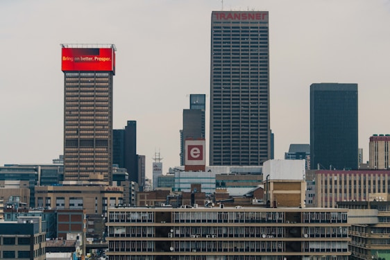 A cityscape features several tall buildings with modern architecture. The forefront shows a prominent building with a red top displaying the text 'Bring on better. Prosper.' Another building visible is labeled 'Transnet' with a logo of the letter 'e.' The skyline is filled with a variety of skyscrapers against a cloudy sky.