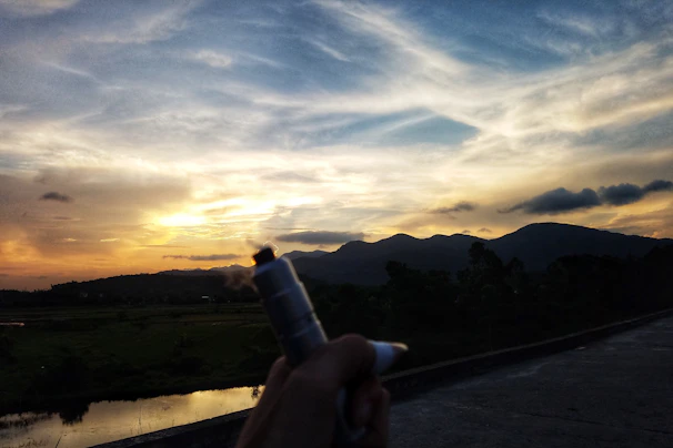 A hand holding a stylish vape pen with the Udaipur city skyline at sunset behind.