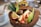 A vibrant fruit bowl served inside a halved coconut shell, featuring slices of banana, strawberries, and green apple, along with a piece of passion fruit. There is granola sprinkled over the fruits and a round, dark-colored snack or truffle in the center. The presentation is complemented by two decorative pineapple leaves. A cup with a spoon is visible in the background on the left.
