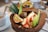 A vibrant fruit bowl served inside a halved coconut shell, featuring slices of banana, strawberries, and green apple, along with a piece of passion fruit. There is granola sprinkled over the fruits and a round, dark-colored snack or truffle in the center. The presentation is complemented by two decorative pineapple leaves. A cup with a spoon is visible in the background on the left.