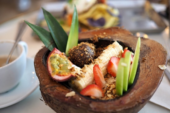 A vibrant fruit bowl served inside a halved coconut shell, featuring slices of banana, strawberries, and green apple, along with a piece of passion fruit. There is granola sprinkled over the fruits and a round, dark-colored snack or truffle in the center. The presentation is complemented by two decorative pineapple leaves. A cup with a spoon is visible in the background on the left.