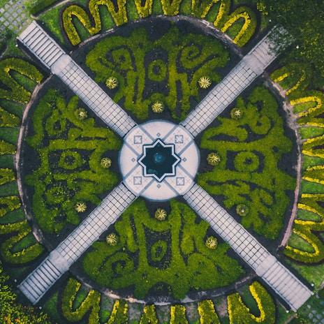 An intricately designed garden with a symmetrical layout, featuring a central circular area with a star-shaped fountain. The surrounding garden is divided into sections by four pathways, each lined with neatly trimmed hedges and manicured plants. The pattern within the greenery appears artistic and well-maintained, with a mixture of light and dark green shades creating a vivid contrast.
