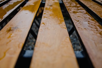 Close-up view of wooden planks with a glossy, wet surface. The planks are arranged in parallel lines, likely forming the seat of a bench, with raindrops scattered across them. The background is blurred, focusing attention on the texture and shine of the wet wood.