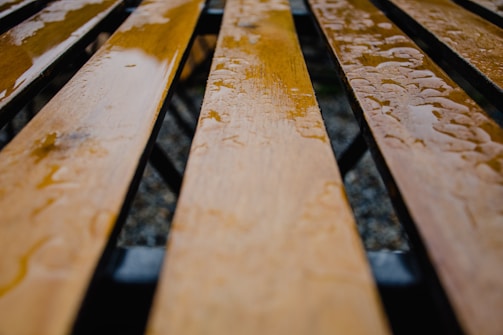 Close-up view of wooden planks with a glossy, wet surface. The planks are arranged in parallel lines, likely forming the seat of a bench, with raindrops scattered across them. The background is blurred, focusing attention on the texture and shine of the wet wood.