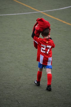 A young boy wearing a red soccer jersey with the number 27 on the back is standing on a grassy field. He is holding a red garment, possibly a jacket or a shirt, over his head. The boy is wearing blue shorts, red socks, and black soccer shoes. The field has yellow lines marking it.