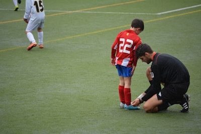 A young soccer player wearing a red and white striped jersey with the number 22 is having his shoelaces tied by an adult dressed in black. The scene takes place on a green soccer field with another player positioned in the background.