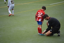 A young soccer player wearing a red and white striped jersey with the number 22 is having his shoelaces tied by an adult dressed in black. The scene takes place on a green soccer field with another player positioned in the background.