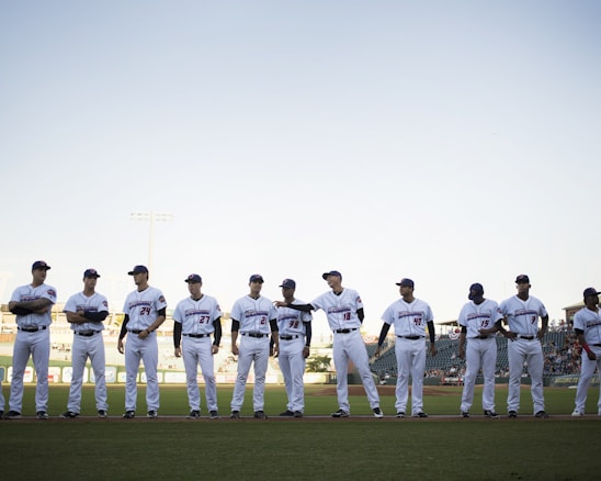 A group of baseball players wearing white uniforms stands in a line on a baseball field. The players are positioned under a clear sky with stadium lights in the background. They appear to be in a pre-game lineup with the grass field visible at their feet.