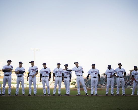 A group of baseball players wearing white uniforms stands in a line on a baseball field. The players are positioned under a clear sky with stadium lights in the background. They appear to be in a pre-game lineup with the grass field visible at their feet.