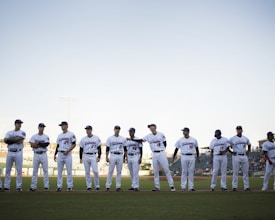 A group of baseball players wearing white uniforms stands in a line on a baseball field. The players are positioned under a clear sky with stadium lights in the background. They appear to be in a pre-game lineup with the grass field visible at their feet.