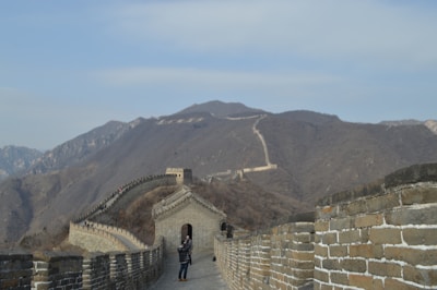 A person walks along a section of the Great Wall of China, surrounded by mountainous terrain. The wall, made of brick, extends into the distance, following the contours of the hills.