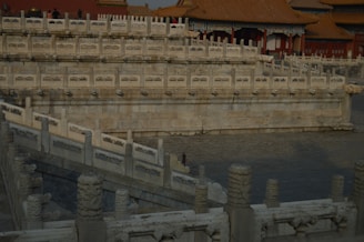 Elaborate stone railings with decorative carvings are prominently featured, surrounding a water body. The architectural style is traditional with ornate elements, and there are several reddish-roofed buildings in the background.