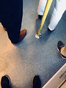 Several pairs of feet are visible on a speckled floor. One person is wearing dark pants with brown shoes, another in white pants and black shoes, and others with sneakers stand nearby. A yellow pole is present, commonly found in public transport.