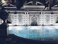 An ornately designed pool is framed by a decorative stone facade featuring intricate patterns and arches. The water reflects soft lights that create a tranquil ambiance. The architecture includes classical elements with columns and detailed stone carvings. Surrounding foliage adds a touch of greenery to the scene, enhancing the luxurious and serene atmosphere.
