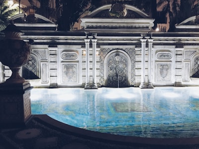 An ornately designed pool is framed by a decorative stone facade featuring intricate patterns and arches. The water reflects soft lights that create a tranquil ambiance. The architecture includes classical elements with columns and detailed stone carvings. Surrounding foliage adds a touch of greenery to the scene, enhancing the luxurious and serene atmosphere.
