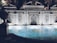 An ornately designed pool is framed by a decorative stone facade featuring intricate patterns and arches. The water reflects soft lights that create a tranquil ambiance. The architecture includes classical elements with columns and detailed stone carvings. Surrounding foliage adds a touch of greenery to the scene, enhancing the luxurious and serene atmosphere.