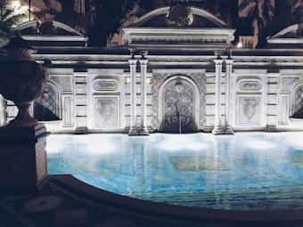 An ornately designed pool is framed by a decorative stone facade featuring intricate patterns and arches. The water reflects soft lights that create a tranquil ambiance. The architecture includes classical elements with columns and detailed stone carvings. Surrounding foliage adds a touch of greenery to the scene, enhancing the luxurious and serene atmosphere.