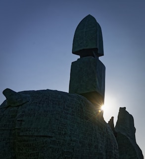 A large stone sculpture is silhouetted against the sky, with sunlight peeking through and highlighting the textures and engraved text on the stone surface. The sculpture appears abstract and monumental.